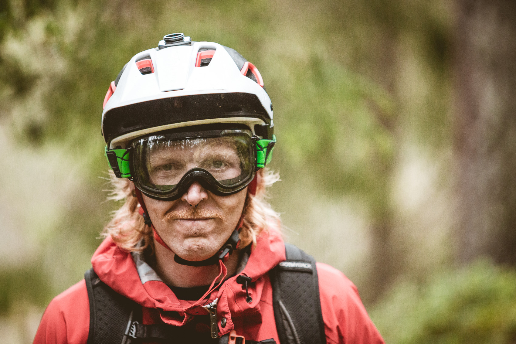 Porträt eines Mountainbikers mit Helm und Brille, der ernst in die Kamera blickt. Er trägt eine rote Jacke und einen Rucksack, im Hintergrund verschwimmt ein Wald.