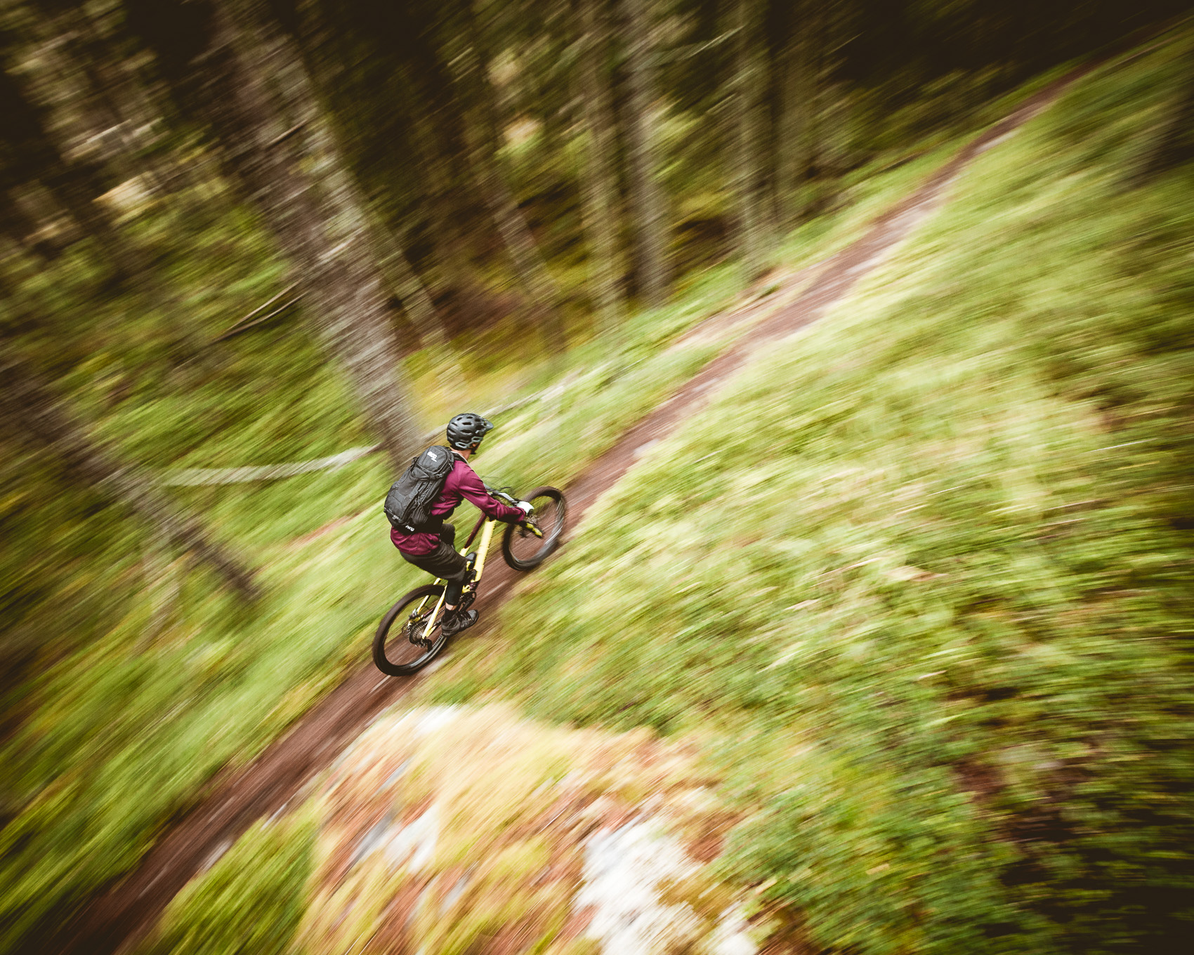 Ein Mountainbiker fährt auf einem schmalen Trail durch einen Wald, aufgenommen mit einer Bewegungsunschärfe, die Geschwindigkeit suggeriert. Der Fahrer trägt Schutzausrüstung und einen Rucksack, während er das Abenteuer auf dem Bike genießt.
