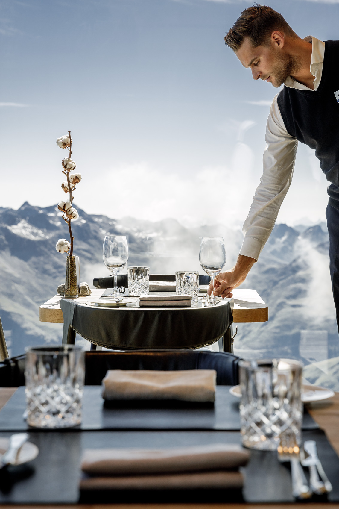 Ein Kellner deckt elegant einen Tisch im Gourmet Restaurant Ice-Q auf dem Gaislachkogel in Sölden, mit einer atemberaubenden Bergkulisse der Ötztaler Alpen im Hintergrund. Dieser exklusive Ort bietet Sterne-Küche auf 3000m Höhe.