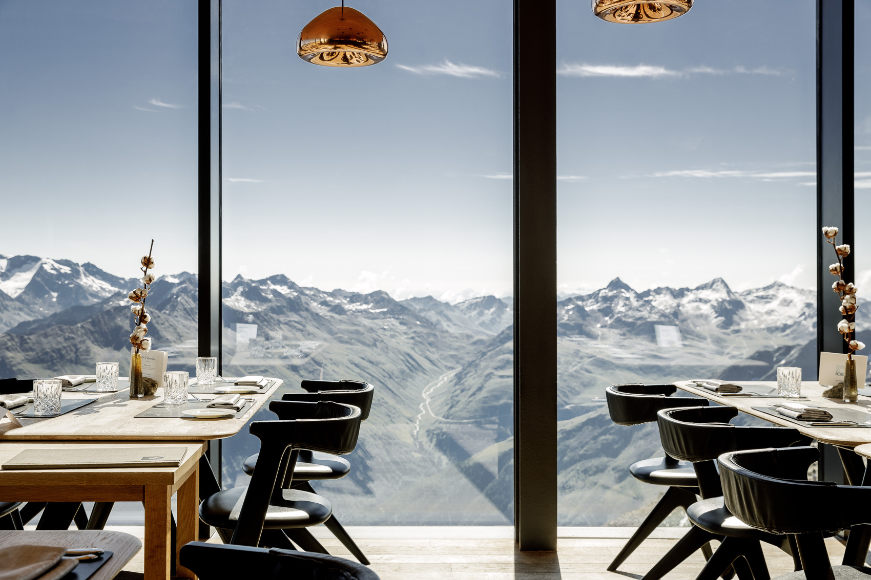 Ein Blick ins Gourmet Restaurant Ice-Q auf dem Gaislachkogel in Sölden, wo Tische mit schwarzem Ledergestühl und einer minimalistischen Dekoration vor einer raumhohen Fensterfront stehen. Im Hintergrund ist eine atemberaubende Panoramaaussicht auf die schneebedeckten Alpen zu sehen, die die exklusive Sterne-Küche auf 3000m Höhe unterstreicht.