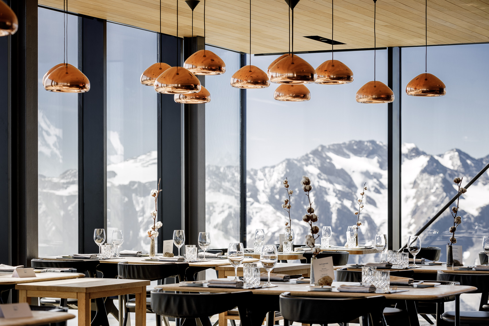 Blick in das Sterne-Restaurant Ice-Q auf dem Gaislachkogel in Sölden, mit gedeckten Tischen, eleganten Kupferlampen und einer atemberaubenden Bergkulisse im Hintergrund. Das luxuriöse Ambiente auf 3000m Höhe lädt zum Gourmet-Erlebnis ein.
