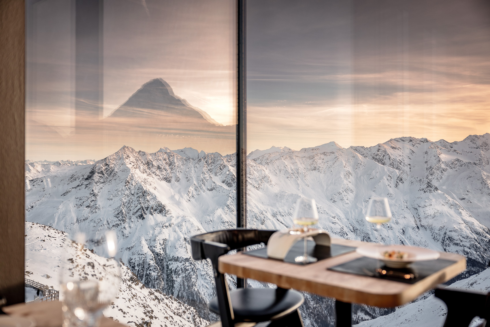 Blick aus dem Gourmet Restaurant Ice-Q am Gaislachkogel in Sölden auf 3000m: Ein elegant gedeckter Tisch mit Weingläsern vor einem atemberaubenden Panorama verschneiter Alpen bei Sonnenuntergang. Im Glas spiegelt sich schemenhaft ein weiterer Berggipfel.