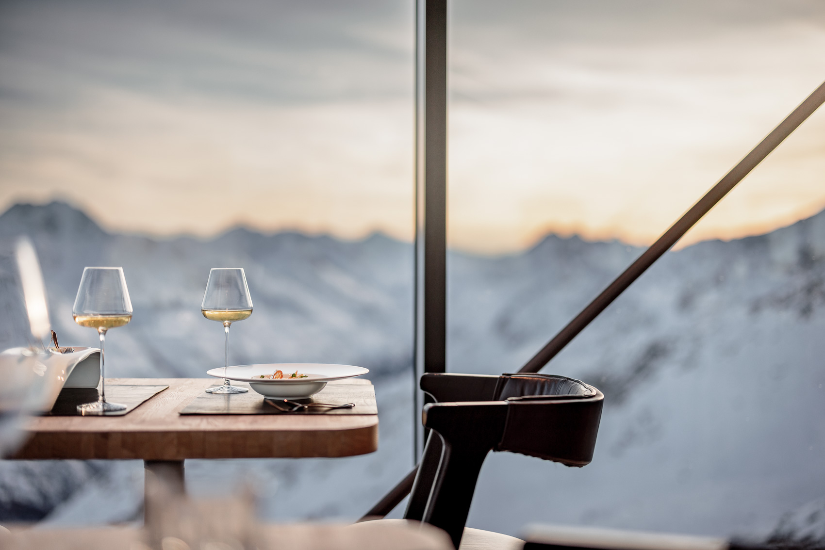 Tischgedeck im Gourmet Restaurant Ice-Q auf dem Gaislachkogel in Sölden mit zwei Weingläsern und einer Speise, vor dem Panorama der schneebedeckten Alpen. Das Bild vermittelt exklusiven Genuss auf 3000m Höhe.