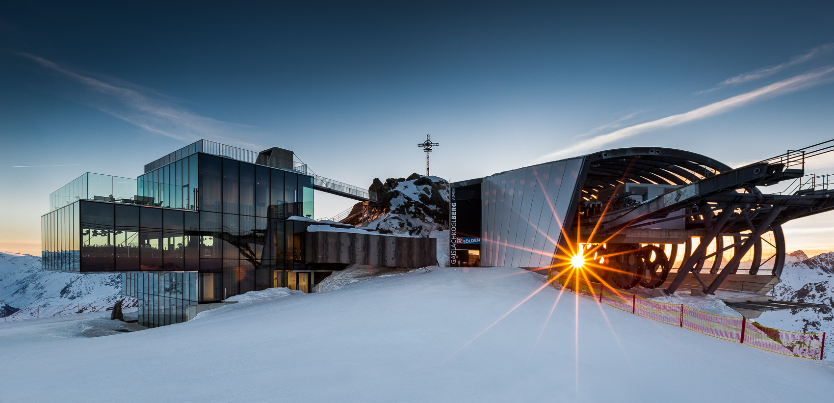 Das Ice-Q Gourmetrestaurant auf dem Gaislachkogel in Sölden, ein moderner Glasbau inmitten verschneiter Berggipfel, wird von der tiefstehenden Sonne angestrahlt. Im Hintergrund ist die Seilbahnstation Gaislachkogelbahn zu sehen, alles umgeben von winterlicher Schneelandschaft.