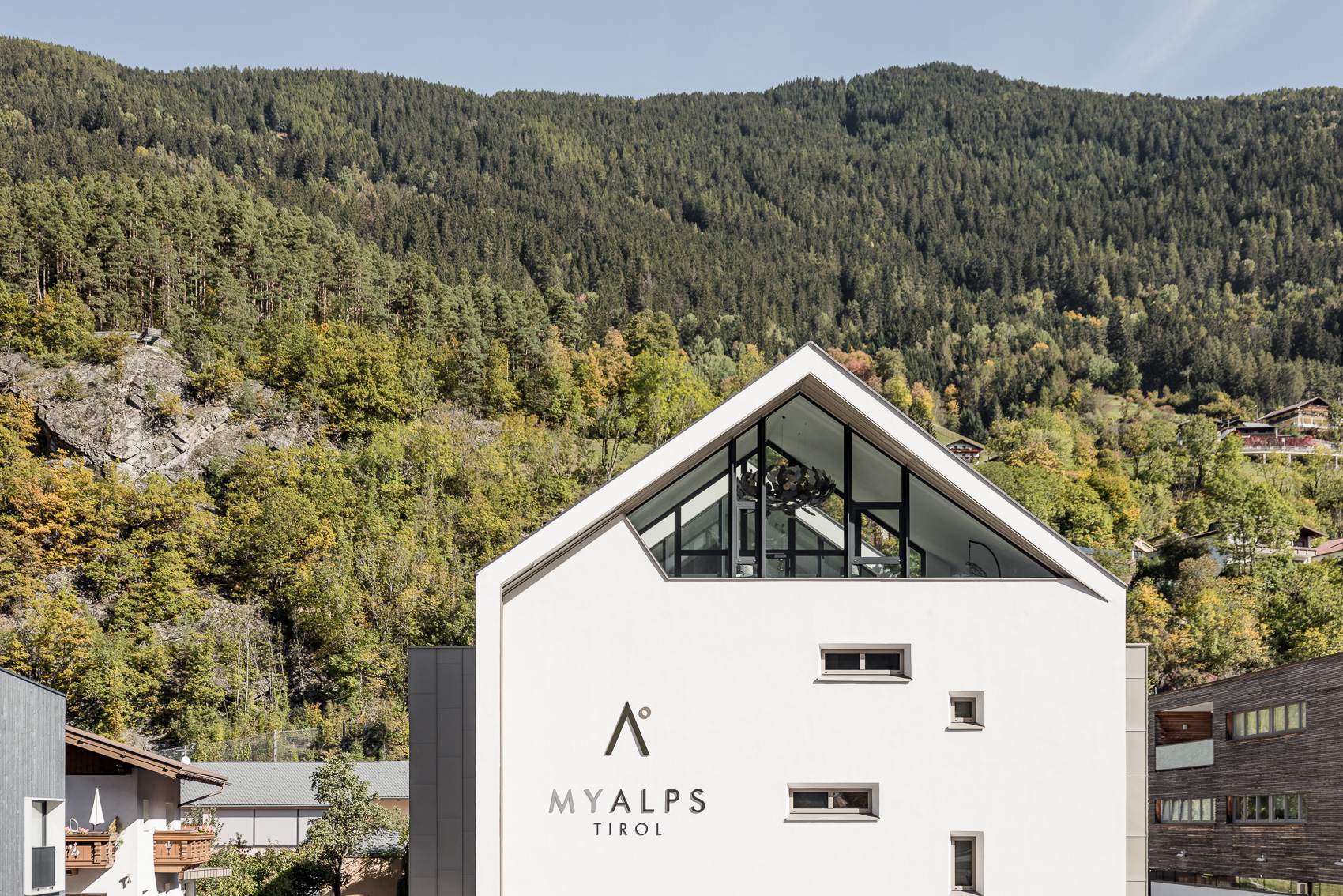 Das moderne MyAlps Tirol Hotel in Oetz zeigt seine charakteristische Giebelarchitektur mit großen Glasflächen. Im Hintergrund erheben sich bewaldete Berghänge, die die alpine Lage unterstreichen.