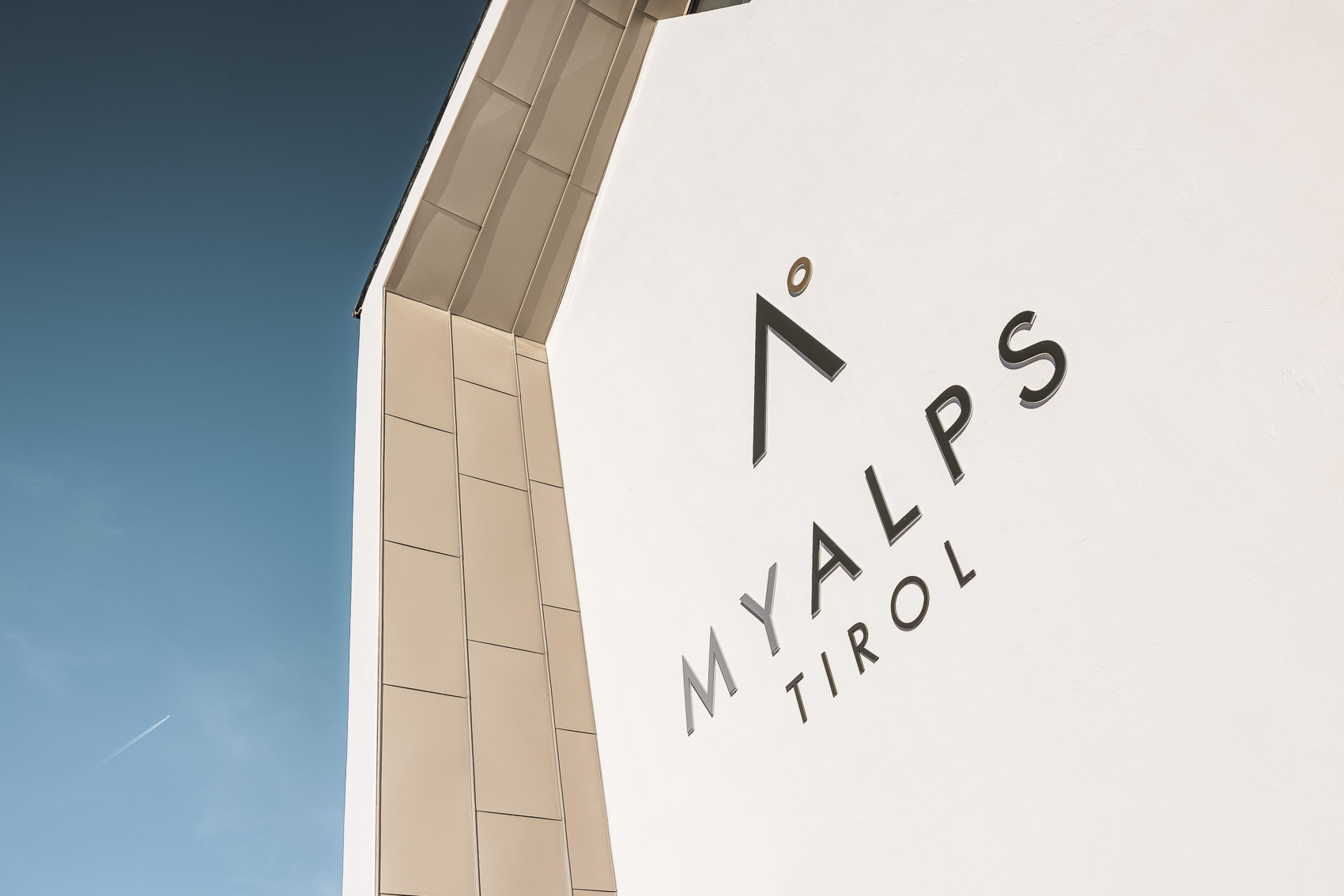 Das Bild zeigt einen modernen Gebäudeteil des Hotels MyAlps Tirol in Oetz mit dem prominent platzierten "MyAlps" Logo auf einer weißen Wand. Im Hintergrund ist ein klarer blauer Himmel mit einem Kondensstreifen zu sehen.