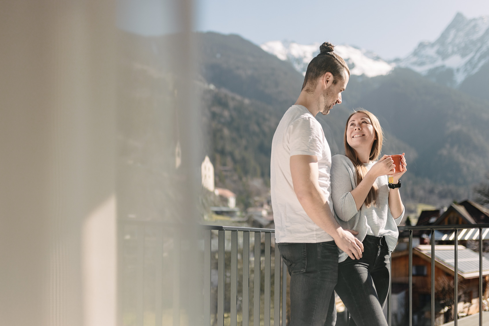 Ein Paar genießt auf einem Balkon den Ausblick auf die verschneiten Berge in Oetz, Tirol, passend zum Thema My Alps. Sie teilen einen Moment der Gemütlichkeit mit einer Tasse Heißgetränk.