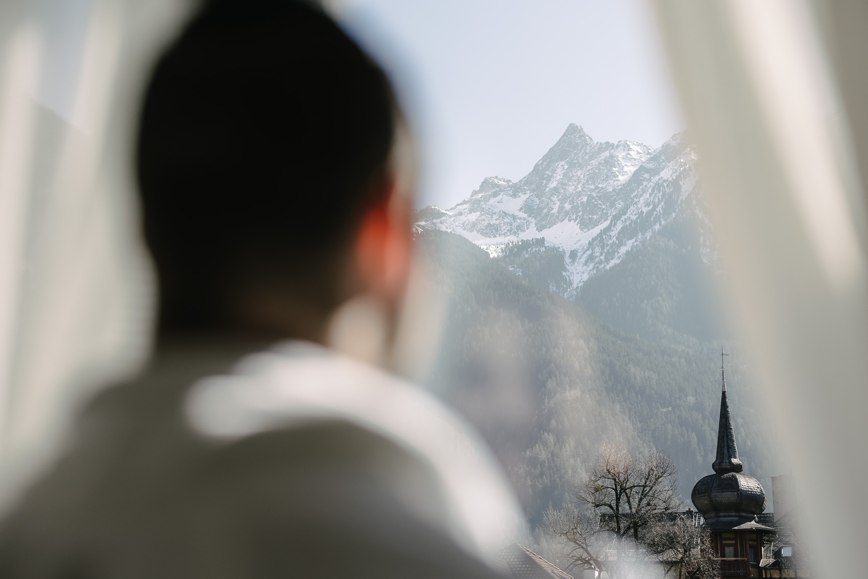 Blick aus einem MyAlps Hotelzimmer in Oetz, Tirol: Eine unscharfe Rückenansicht einer Person schaut auf ein schneebedecktes Alpenpanorama und einen Kirchturm. Diese Hotelfotografie unterstreicht die beeindruckende Bergkulisse.