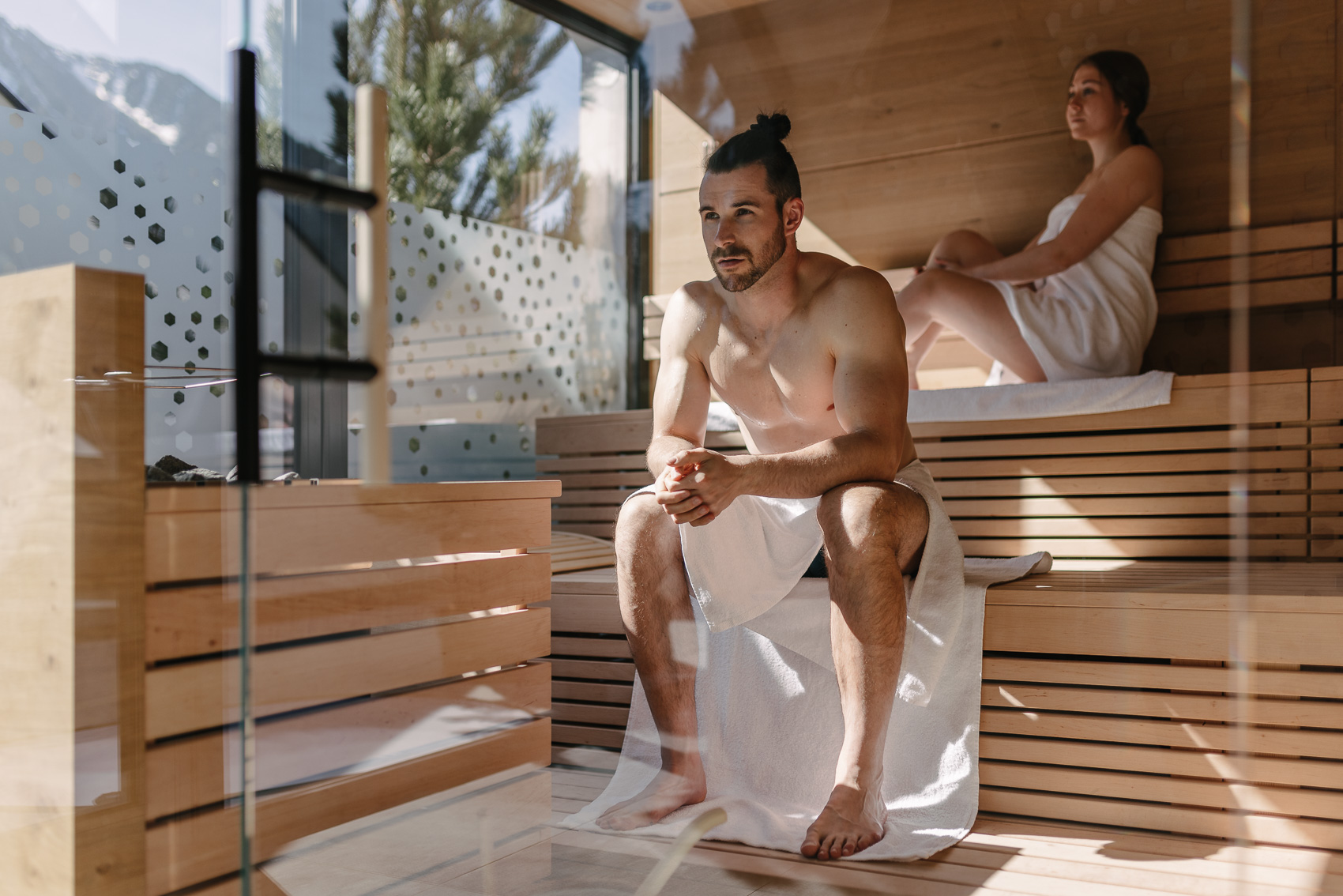 Zwei Personen entspannen in einer modernen Sauna mit Bergblick, fotografiert für MyAlps Tirol im Rahmen der Hotelfotografie in Oetz. Der Mann sitzt im Vordergrund, die Frau dahinter, beide in weiße Handtücher gewickelt.