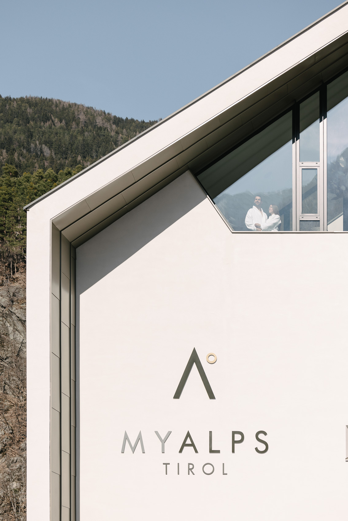 Ein modernes Gebäude des MyAlps Tirol Hotels in Oetz mit seinem Logo auf der Fassade. Im oberen Stockwerk blickt ein Paar in Bademänteln durch ein großes Fenster auf die dahinterliegende Berglandschaft.