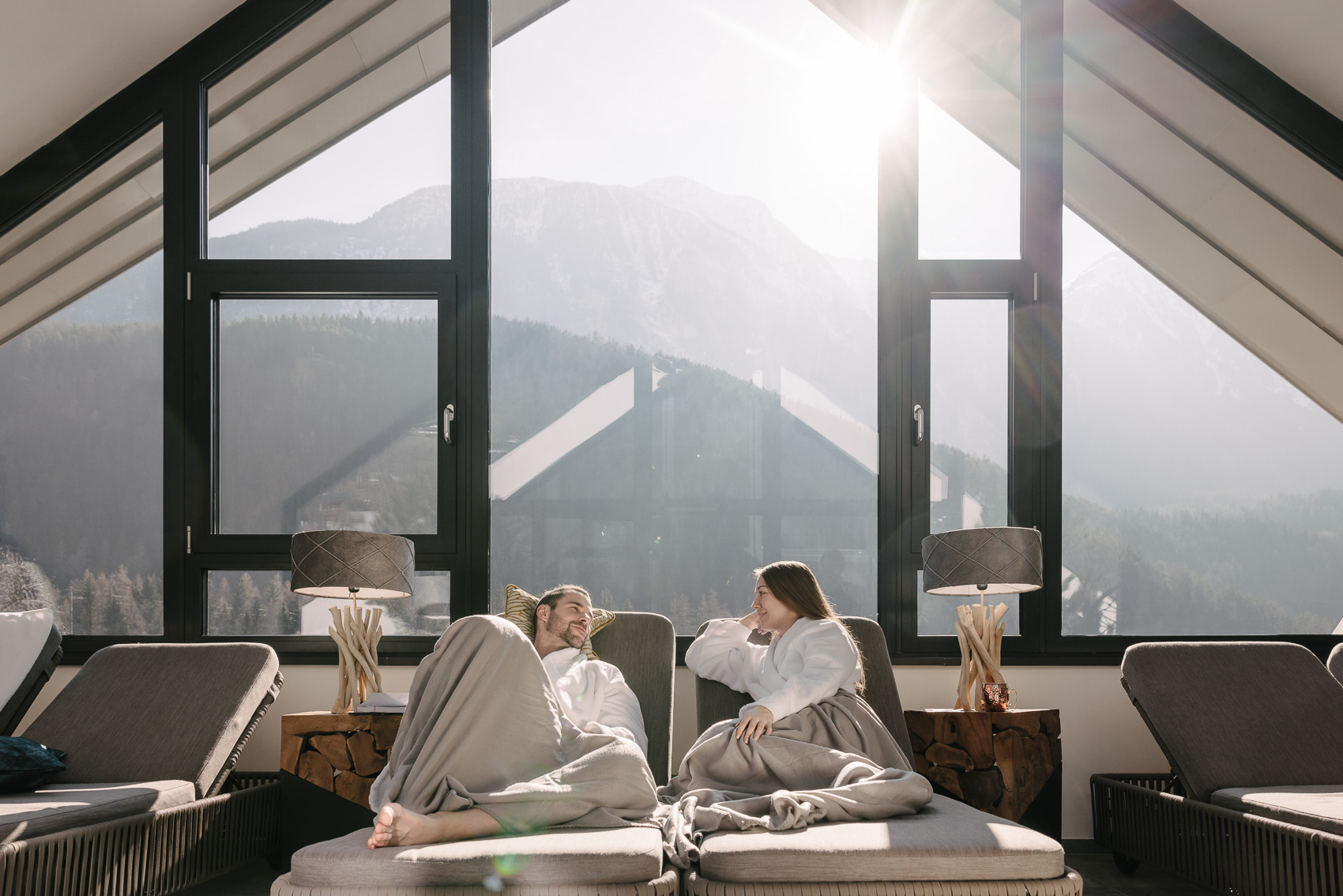 Ein Paar entspannt in Bademänteln auf Liegen und genießt den weiten Bergblick aus einem Fenster im MyAlps Tirol Hotel in Oetz. Dieses Bild der Hotelfotografie fängt die Gemütlichkeit und Alpenkulisse ein.