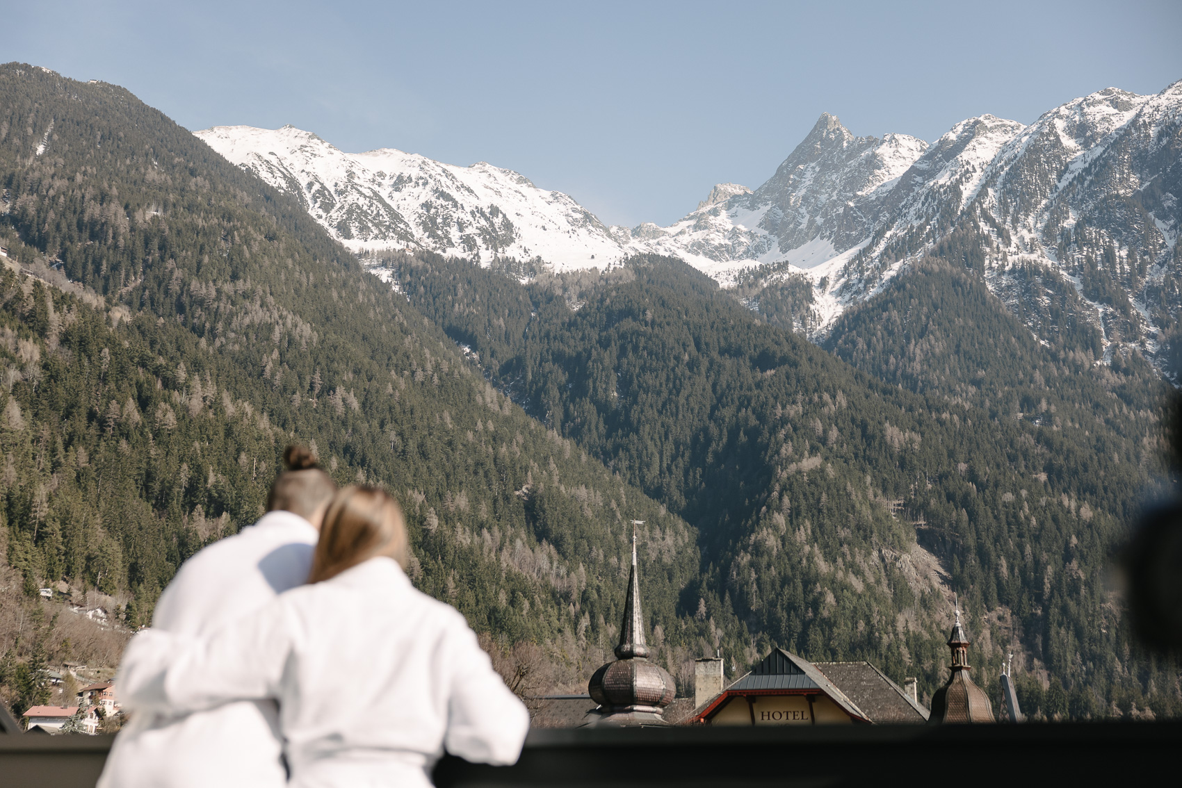 Zwei Personen in Bademänteln blicken auf die schneebedeckten Berge und grünen Wälder in Oetz, Tirol, was die entspannende Atmosphäre des Hotels unterstreicht. Im Vordergrund ist das Dach eines Hotels sichtbar, passend zur Hotelfotografie für My Alps / MyAlps Tirol.