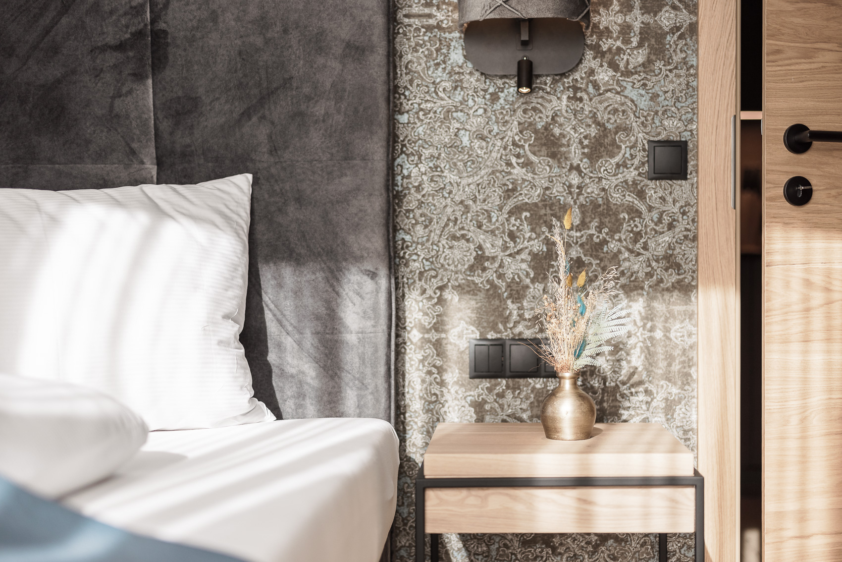 Detailaufnahme eines stilvollen Hotelzimmers in Oetz, MyAlps Tirol, mit einem bequemen Bett und Nachttisch, der einen kleinen Trockenblumenstrauß trägt. Die Hotelfotografie zeigt das moderne Design und die gemütliche Atmosphäre des Zimmers.