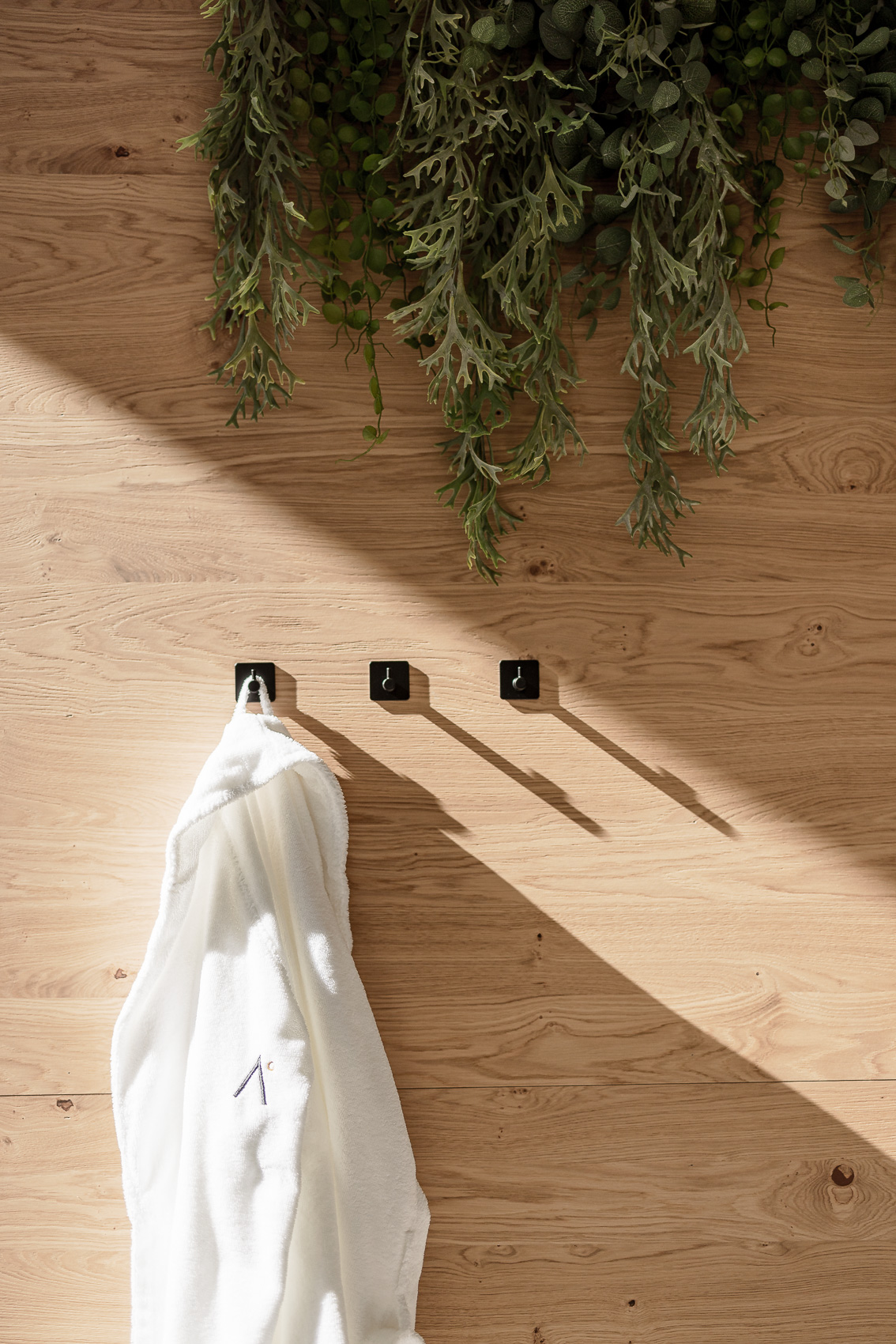 Holzwand mit grünen Hängepflanzen und drei schwarzen Haken, an denen ein weißer Bademantel mit dem MyAlps-Logo für die Hotelfotografie in Oetz, Tirol, hängt. Sonnenlicht fällt schräg auf die Szene und wirft lange Schatten.